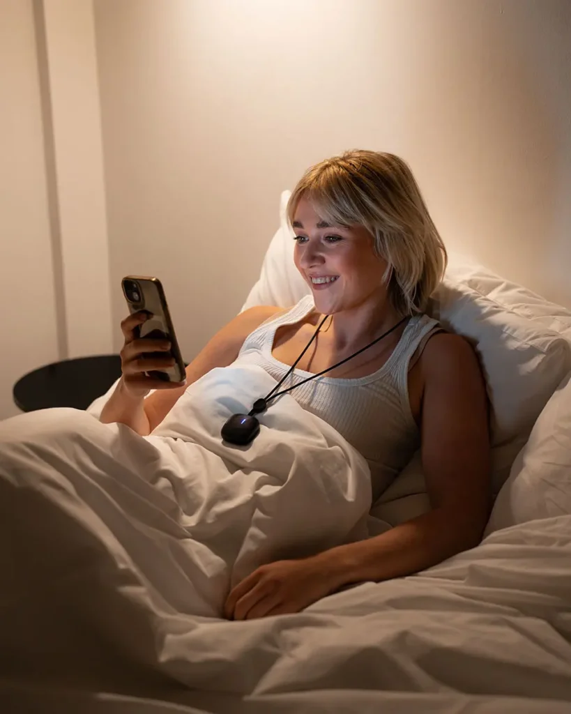 Woman relaxing in bed, wearing the Serenity Comfort neck massager, enjoying a moment of calm with her smartphone.